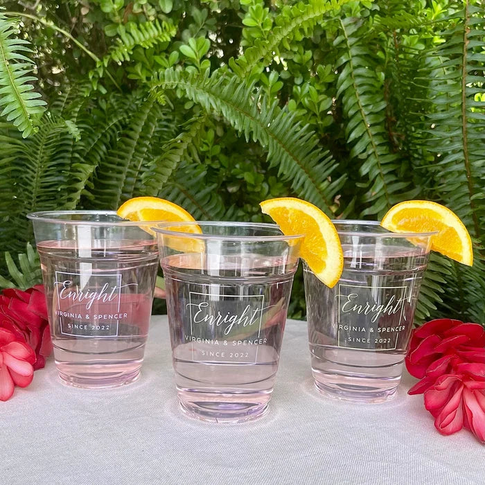 Three clear plastic cups filled with water, each with a slice of orange on top, placed on a table with red flowers in the background.