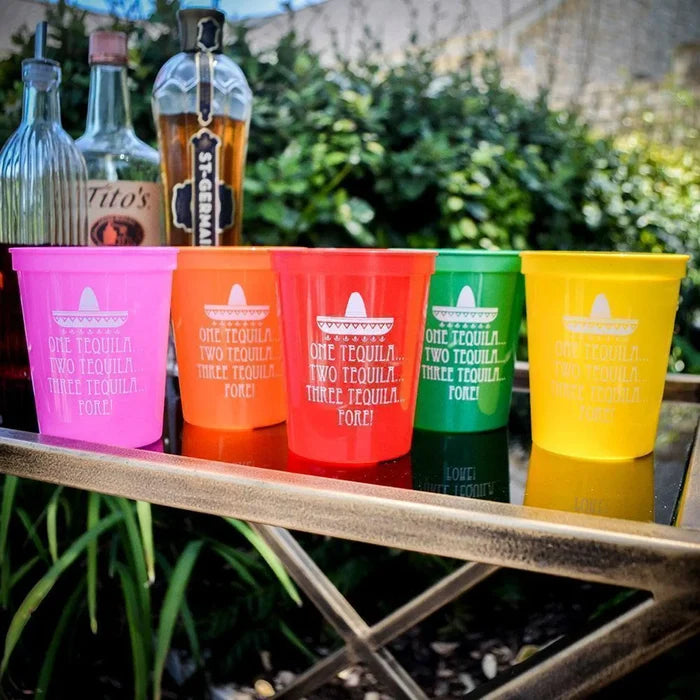 A collection of colorful plastic cups with the text "ONE TEQUILA" printed on them, along with a bottle of tequila and other bottles in the background.