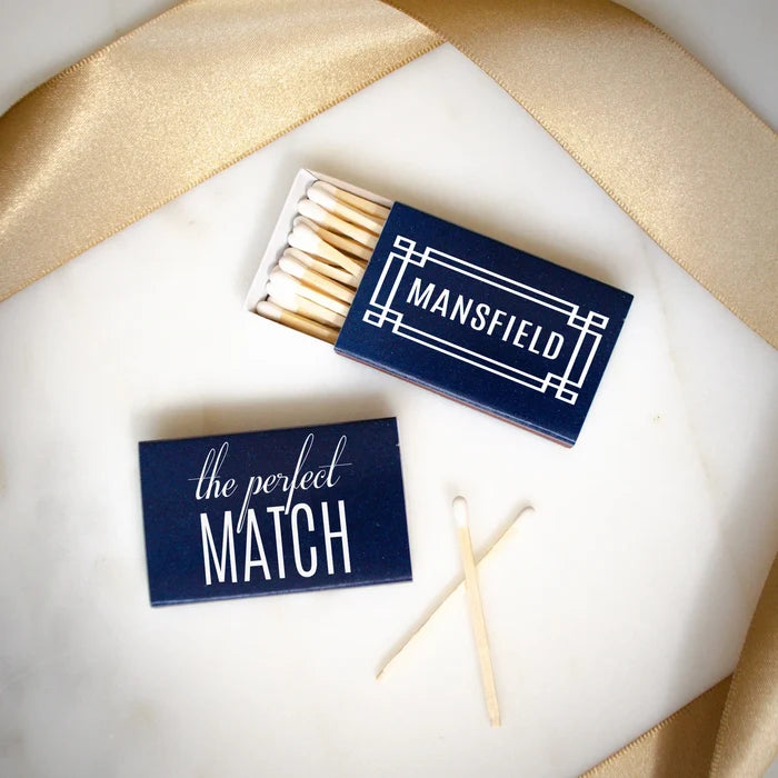 a pair of matchsticks and a matchbox with the word "MANSFIELD" printed on it, placed on a white surface with a gold ribbon.