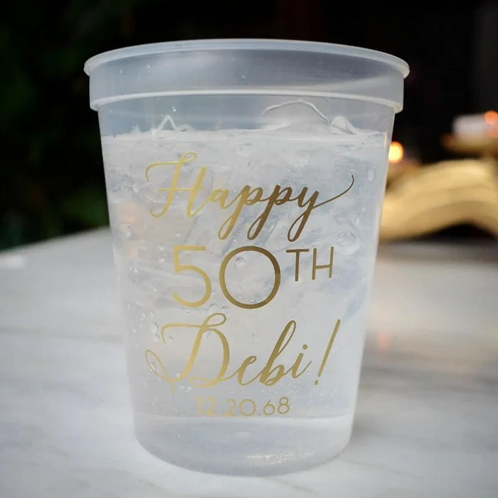 A clear plastic cup filled with ice and water, with the text "Happy 50th Debi!" printed on it.