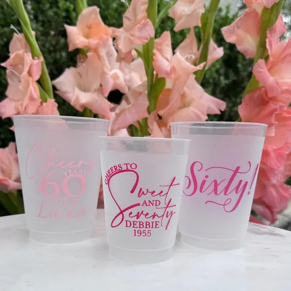 Three clear plastic cups with personalized text, placed in front of pink flowers.