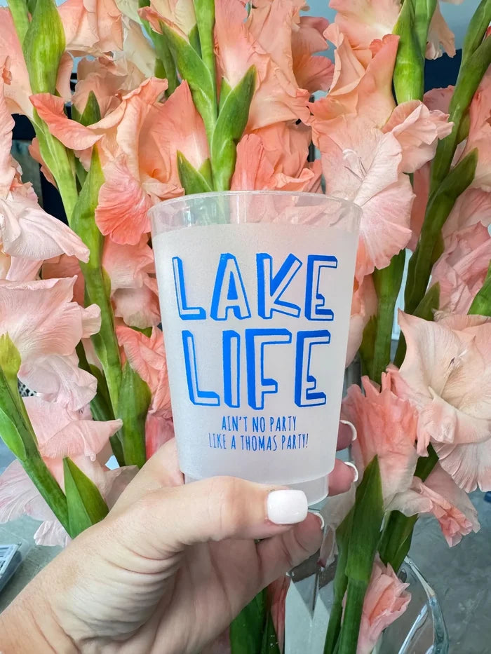 A person's hand holding a clear cup with the text "LAKELIFE AIN'T NO PARTY LIKE A THOMAS PARTY!" printed on it, set against a background of pink flowers.